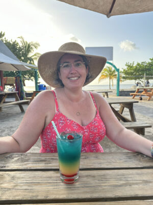 A photo of me in a pink summery dress and wide-brimmed hat with a pint glass in front of me that is layered with ingredients so it's red on bottom, then yellow, green, and blue.